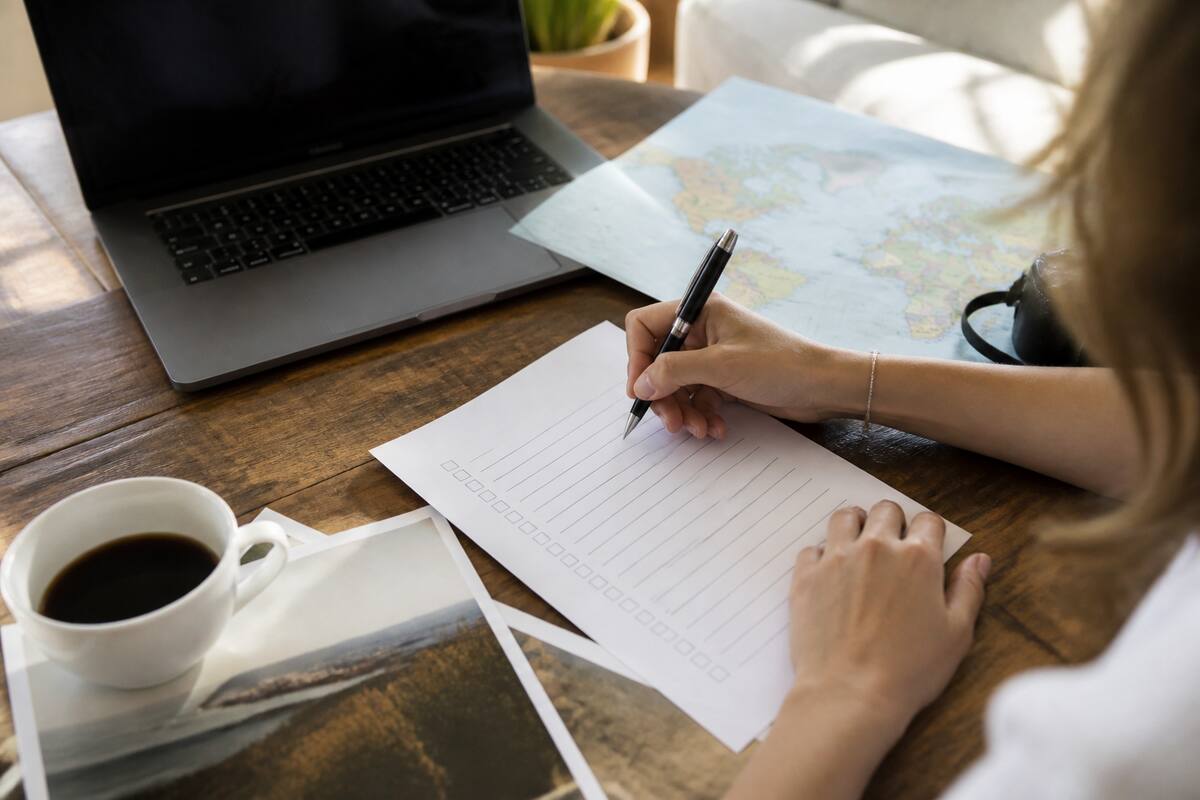 Persoon gebruikt een vakantiechecklist met laptop en reisdocumenten op tafel bij daglicht